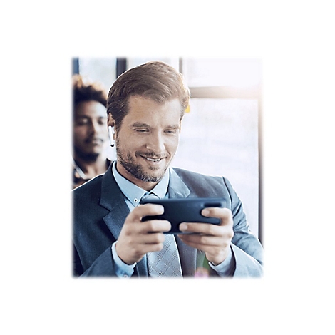 Ein lächelnder Mann im Anzug, mit kabellosen Kopfhörern, blickt auf sein Smartphone. Eine Person ist im Hintergrund unscharf.