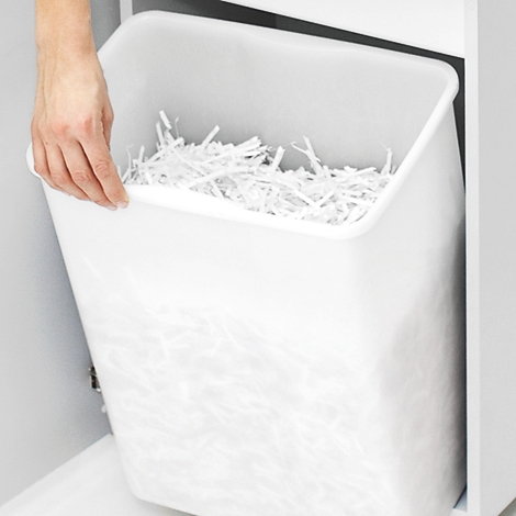 Eine Hand hält einen weißen Papierkorb, gefüllt mit geschredderten Papieren. Der Papierkorb ist in einen Schrank integriert.