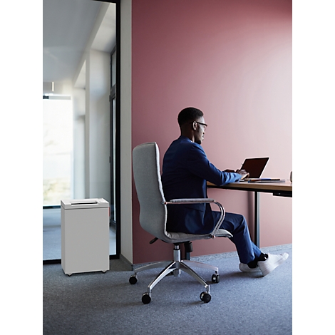 A man in a suit sits at a desk typing on a laptop. A paper shredder stands next to him. Red wall background.