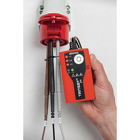 Un appareil de test TRITEST Pro rouge est tenu à la main tout en étant connecté à une prise rouge. Des câbles sont attachés.