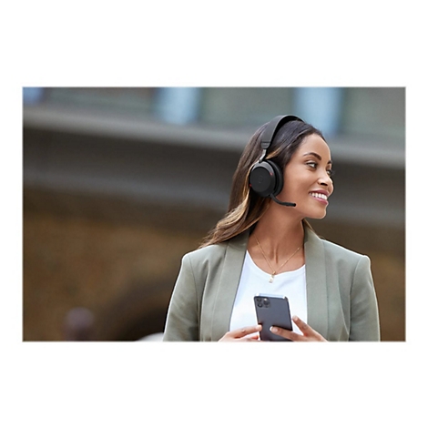 Eine lächelnde Frau mit schwarzem Headset hält ein Smartphone, im Hintergrund Gebäude.
