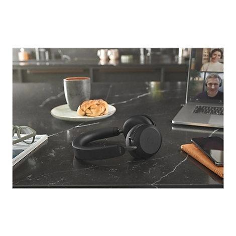 Casque Jabra noir sur un comptoir noir. Arrière-plan : ordinateur portable avec visioconférence, café, pâtisserie.