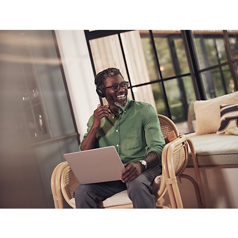 Ein lächelnder Mann sitzt mit Laptop und Kopfhörern in einem Rattansessel, im Hintergrund Fenster und Kissen.