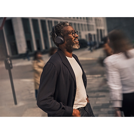 Un homme souriant avec un casque traverse une rue. Il porte des lunettes, une veste et une chemise blanche.