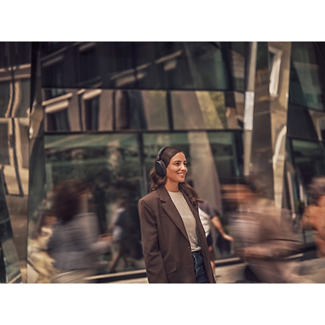 Une femme souriante portant un casque marche dans une ville. Elle porte un blazer marron et regarde vers la droite.