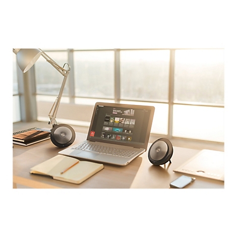 Un ordinateur portable avec un Ã©cran ouvert, deux haut-parleurs et une lampe de bureau sur un bureau. La lumiÃ¨re du soleil brille Ã  travers la fenÃªtre.