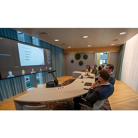 Bürokonferenzraum mit Personen an einem geschwungenen Tisch während einer Videokonferenz auf einem großen Bildschirm.
