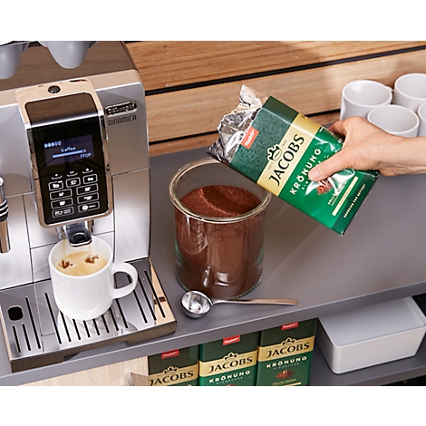 Coffee machine brewing coffee, next to it a hand pouring coffee powder into a glass jar. The bag reads "Jacobs Krönung".