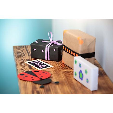 Trois boîtes cadeaux sont posées sur une table en bois. Une boîte noire avec des pois violets et un ruban. Une boîte marron avec une bande noire. Une boîte blanche avec des fleurs bleues.
