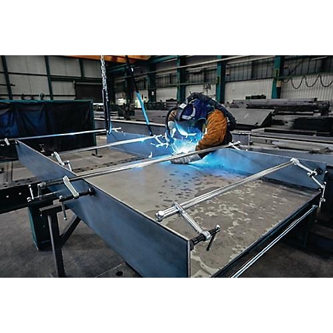 Un ouvrier soude des pièces métalliques dans une usine. Des étincelles bleues et des lunettes de protection sont visibles.