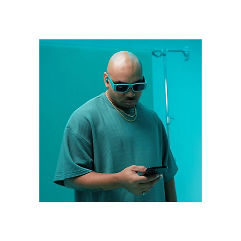 Homme avec des lunettes et des écouteurs regarde un smartphone. Il porte un collier et un t-shirt vert, devant un fond turquoise.