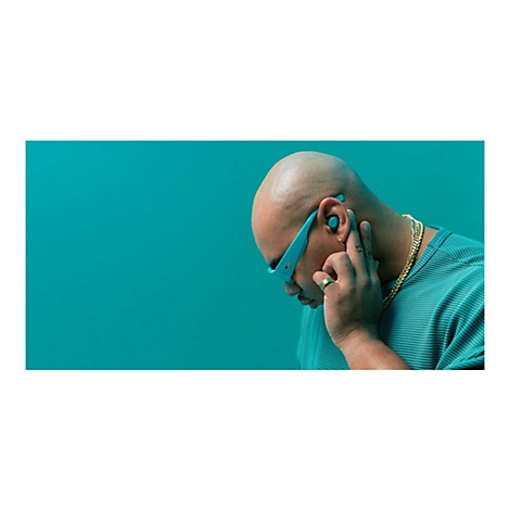 Un homme chauve portant un casque et des lunettes de soleil bleus touche son oreille, fond turquoise.