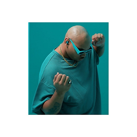 Homme portant des lunettes de soleil, des écouteurs et une chaîne en or ; vêtements verts, en pose.