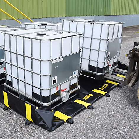 Verschillende witte IBC-containers op zwarte opvangbakken met gele accenten, op een geasfalteerd oppervlak.