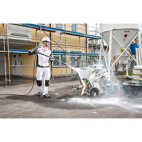 A worker in protective helmet and work clothes operates a plastering machine. A scaffolding and a cement silo are visible in the background.
