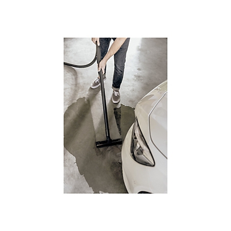 Person reinigt mit einem Nasssauger den Boden neben einem weißen Auto in einer Werkstatt.