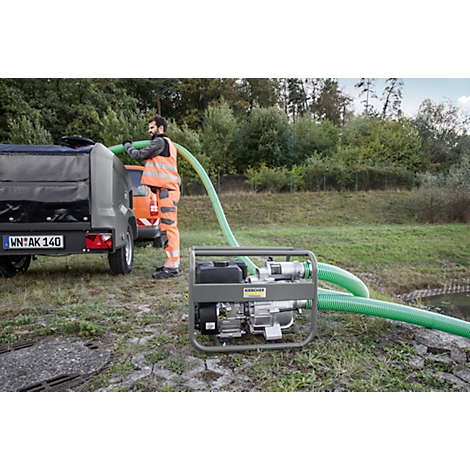 Man met pomp, trekt groene slang uit aanhanger, draagt veiligheidsvest, bomen op de achtergrond.