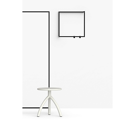 Lichtgrijze kruk met drie poten voor een witte muur met zwart frame en plank.
