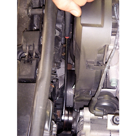 Nahaufnahme des Motorraums eines Autos. Riemen, Rollen und Bauteile sind sichtbar, eine Hand deutet darauf.