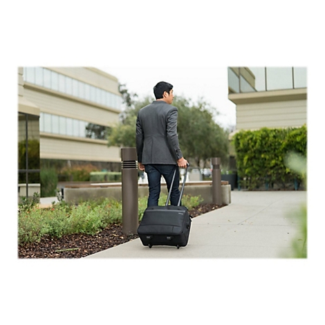 Ein Mann in einem Anzug zieht einen schwarzen Rollkoffer auf einem BÃ¼rgersteig. Im Hintergrund sind GebÃ¤ude und GrÃ¼nflÃ¤chen zu sehen.