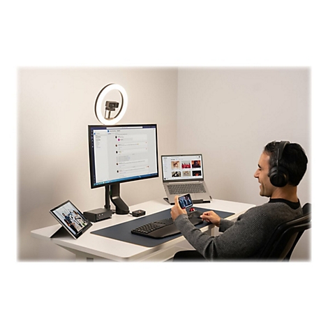 Un homme est assis Ã  un bureau, regarde son smartphone et sourit. Sur le bureau se trouvent un moniteur, un ordinateur portable, une tablette, un casque, une webcam et un clavier.