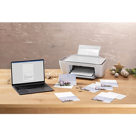 Witte printer met laptop en wenskaarten op houten tafel. De laptop toont een document, kaarten liggen ernaast.