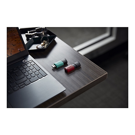Deux clés USB sont posées sur une surface en bois à côté d'un ordinateur portable ouvert. L'ordinateur portable est noir, une clé USB est turquoise, l'autre est rouge. En arrière-plan, on peut voir un porte-clés et une fenêtre.