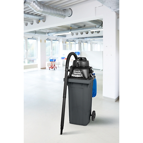 Aspirateur industriel noir sur conteneur à déchets gris, devant un bureau lumineux. L'aspirateur porte l'inscription Mammut.