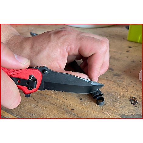 Nahaufnahme einer Hand, die ein rotes Taschenmesser benutzt, um einen schwarzen Ring zu schneiden. Der Hintergrund ist unscharf.