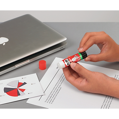 Person hält einen Pritt Klebestift über Papier, daneben ein Laptop, Diagramm und Deckel.