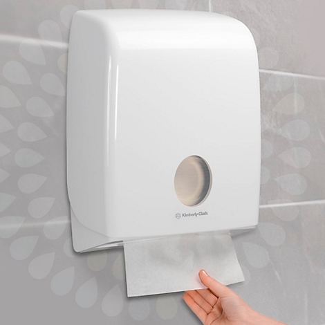 Dispensador de toallas de papel blanco en una pared gris. Una mano saca una toalla de papel.