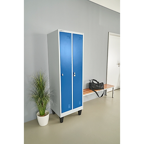 Blauer Spind vor grauer Wand. Daneben eine Pflanze in einem Topf, eine Bank mit Tasche.