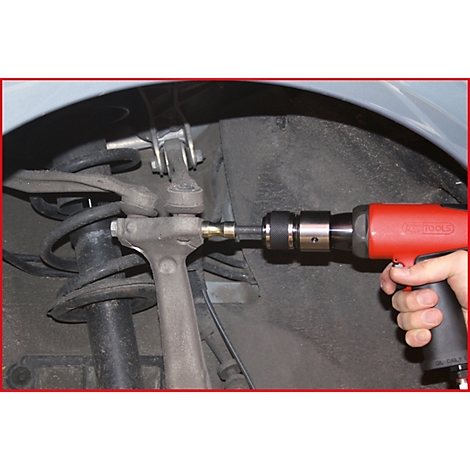 Nahaufnahme eines Mechanikers, der mit einer roten Druckluftbohrmaschine an einem Fahrzeug arbeitet. Der Fokus liegt auf dem Federbein.
