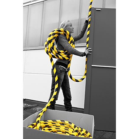 A young woman holds a black and yellow striped barrier. She stands in front of a building with a gray background and a box with the barrier.