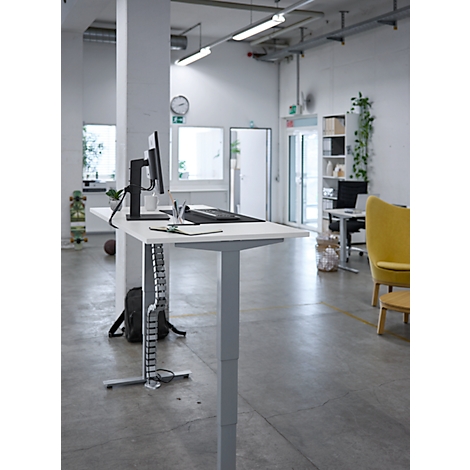 Ein höhenverstellbarer Schreibtisch in hellem Büro. Darauf Monitor, Tastatur und Kabel. Im Hintergrund ein gelber Sessel.