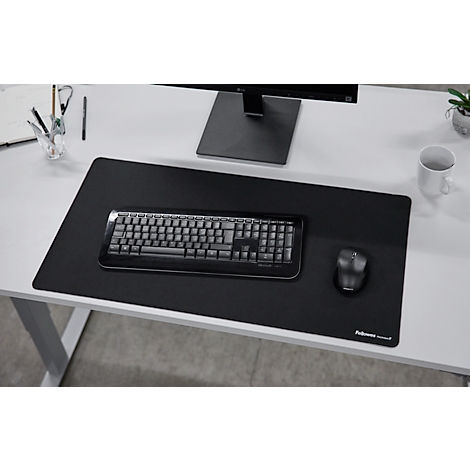 Schwarze Schreibtischunterlage mit Tastatur, Maus, Monitor und Becher auf weißem Schreibtisch.