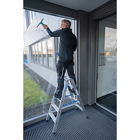 Een man staat op een ladder en maakt een groot raam schoon met een wisser. Hij draagt werkkleding.