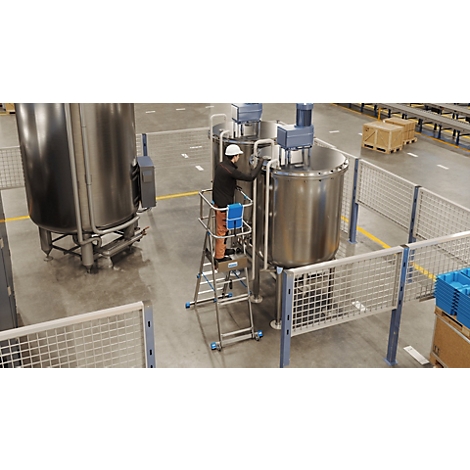 Un travailleur sur une échelle dans une usine entretient une grande machine à mélanger en acier inoxydable. Des grilles et des boîtes sont visibles à droite.