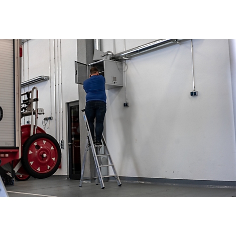 Een man staat op een ladder en werkt aan een open elektrische kast aan de muur. Naast hem is een brandweerwagen te zien.