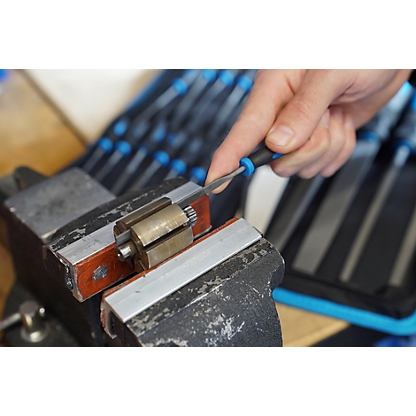 Hand bearbeitet mit einer kleinen Feile ein metallisches Werkstück, das in einem Schraubstock eingespannt ist.