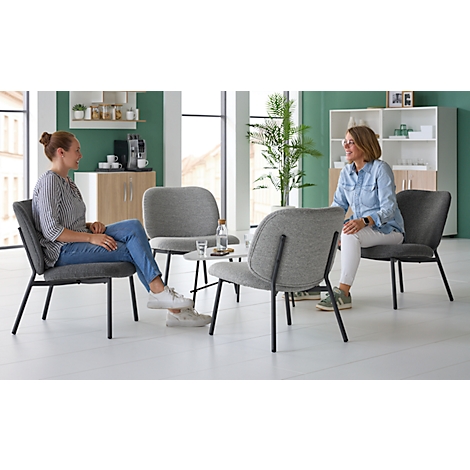 Twee vrouwen zitten in grijze stoelen en praten in een lichte kamer. Op de achtergrond een tafel met water en een kast.