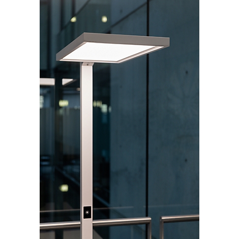 Lampadaire moderne avec un abat-jour carré et lumineux. Mât argenté, fond gris.