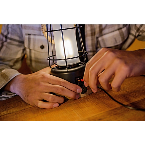 Un homme portant une chemise à carreaux allume une lanterne à l'aide d'un bouton de réglage, assis à une table en bois.