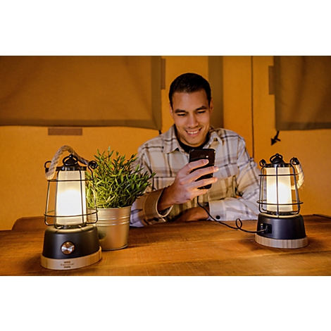 Un jeune homme en chemise à carreaux est assis à une table, souriant et regardant son smartphone, entouré de deux lanternes et d'une petite plante.
