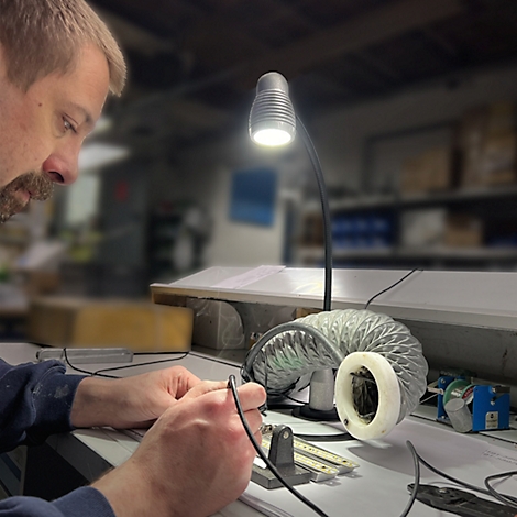 Un homme travaille à un bureau sous une lampe, tenant des outils et travaillant sur une petite pièce métallique.