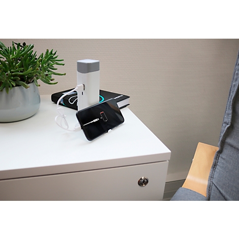 Un smartphone se charge sur une table de chevet blanche. A côté, un chargeur, un livre et une plante. En arrière-plan : une chaise.