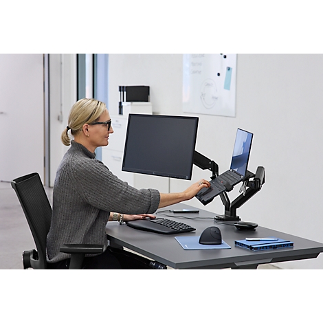 Frau arbeitet am Schreibtisch mit Laptop und Monitor. Sie trägt Brille und einen grauen Pullover.