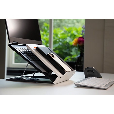 Laptop op witte standaard, naast smartphone en muis, raam met groene planten op de achtergrond.