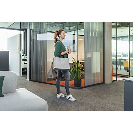 Eine Frau in Jeans und grünem Shirt mit Tasche und Becher geht in einem Büro. Grauer Teppich, Glastüren.