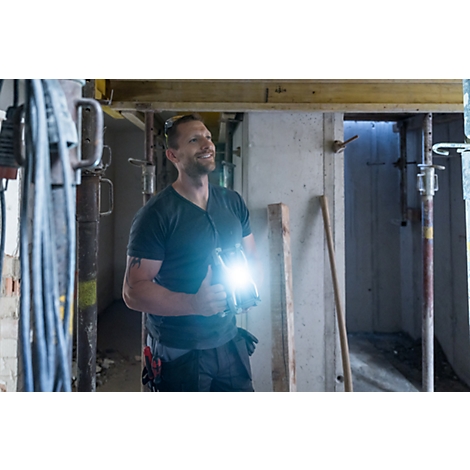 Ein Handwerker lächelt und hält eine Taschenlampe, während er in einem Baustellenbereich steht. Licht strahlt von der Lampe.